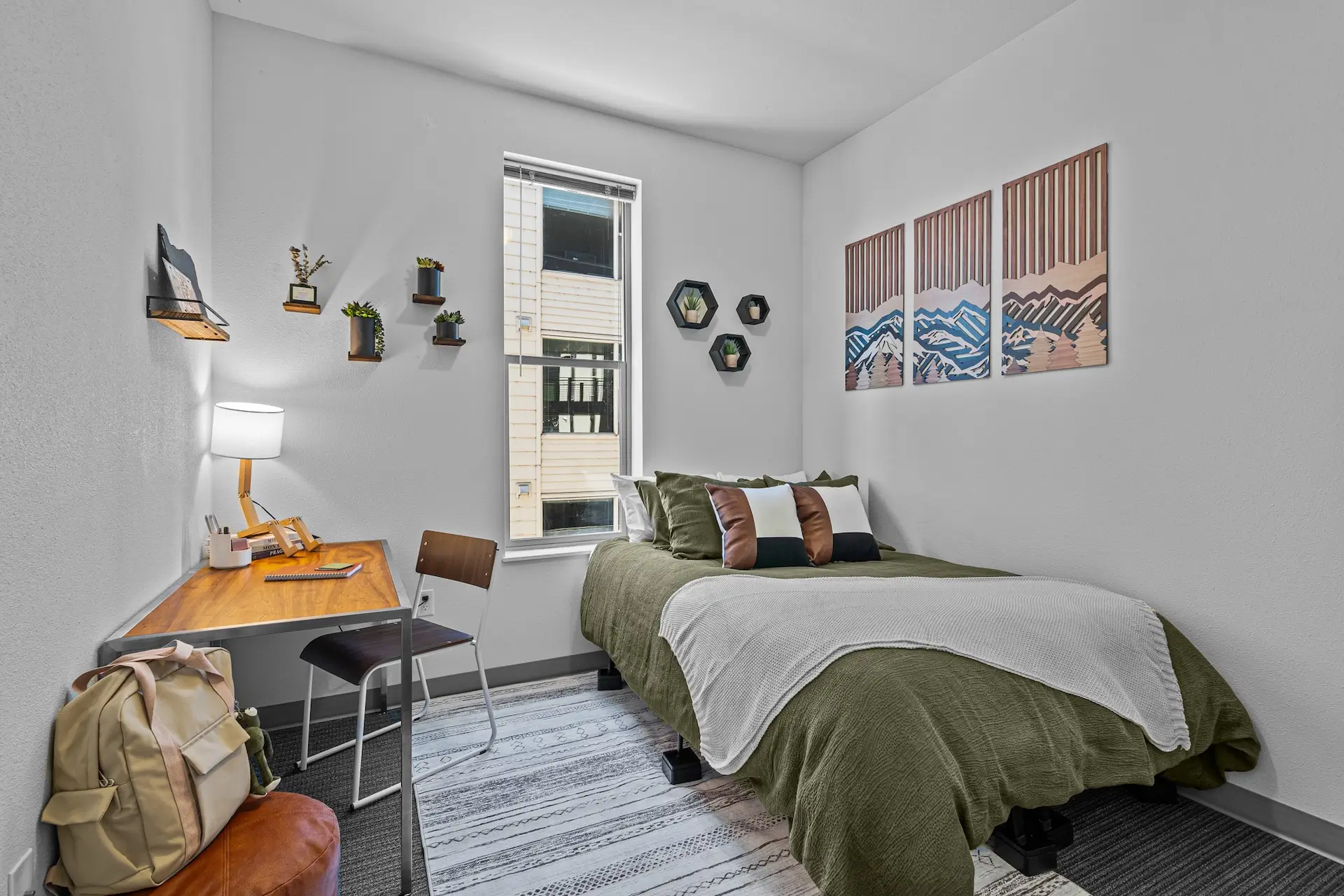 carpeted bedroom with a steel and wood table on left site, a leather cushioned stool, and a bed on the right side under a tall window.